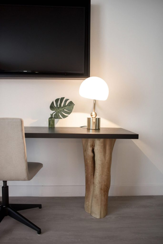 Stainless Steel Base White Shade Table Lamp on Brown Wooden Desk Near White Painted Wall With Wall Mounted Flat Screen T V